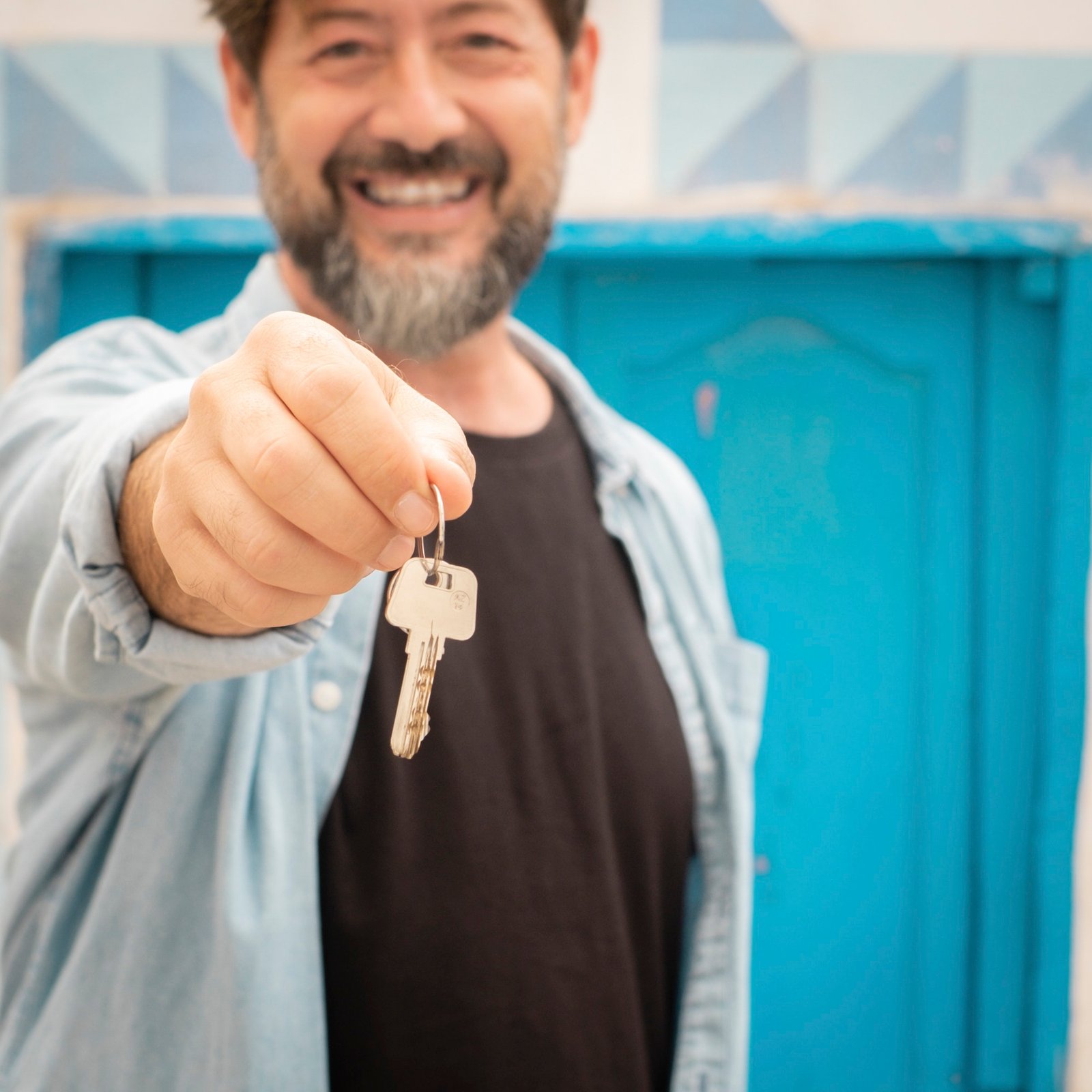 Adult happy man holding home keys and blue door house in background. Concept of buyer